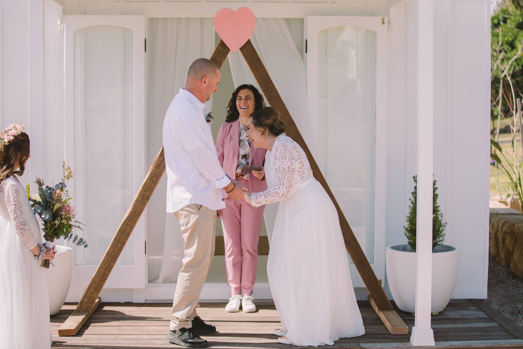 A couple holding hands under a white timber arch topped with a pink heart